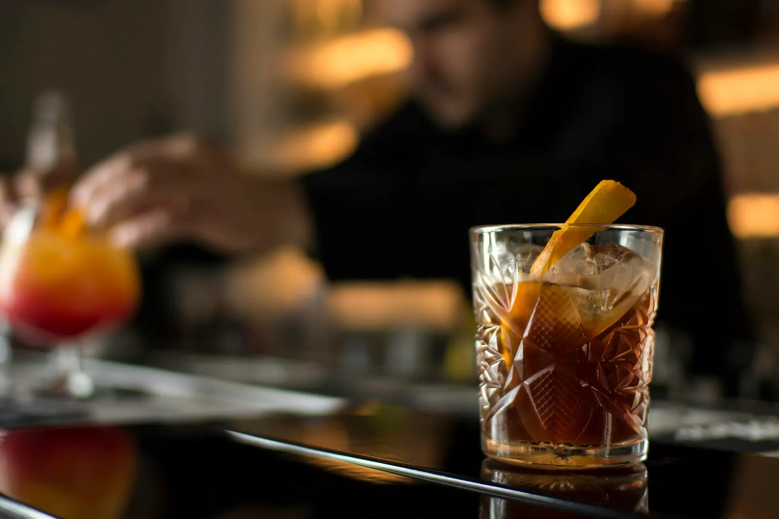 Drink com gelo e laranja no balcão, bartender ao fundo